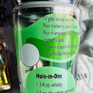 May include: A clear cocktail shaker with a black lid. The glass body features a green label with a golf ball design and cocktail recipes, including "Hole-in-One." The shaker is designed for mixing drinks.