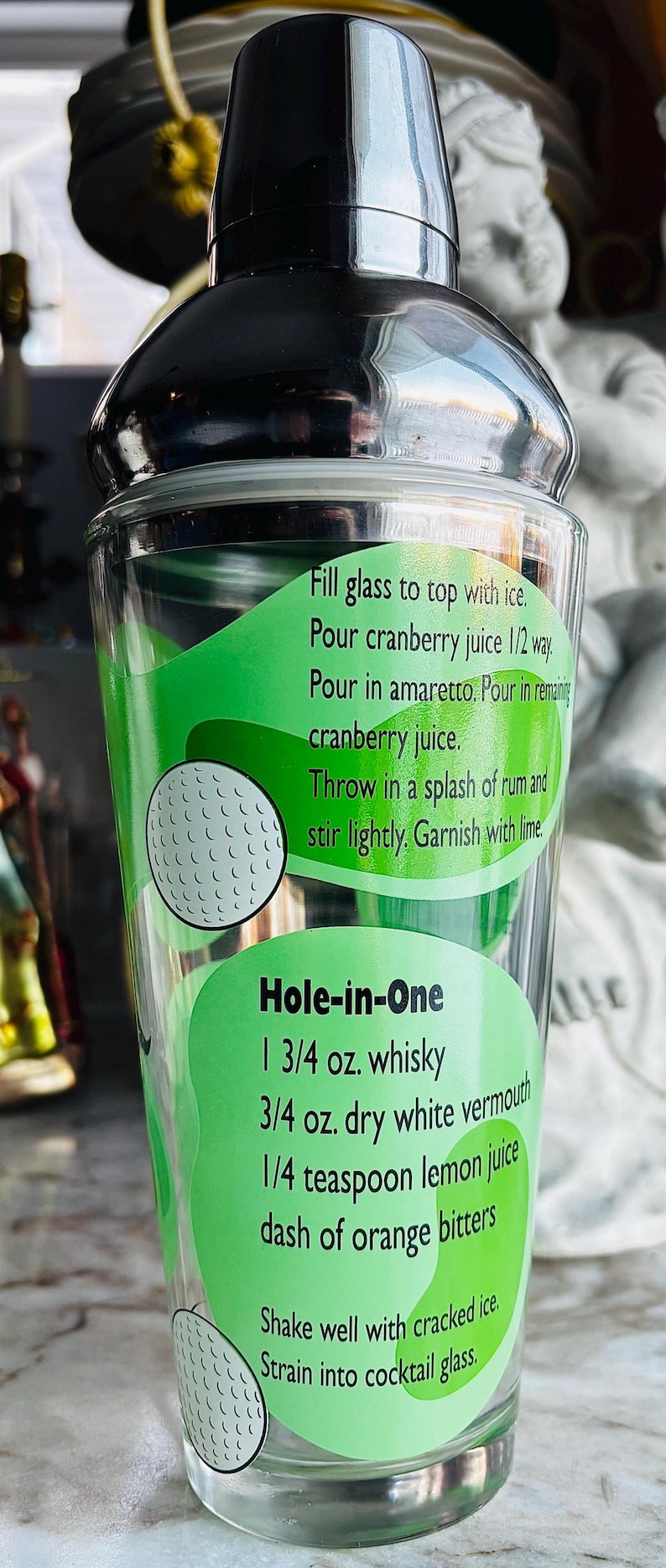 May include: A clear cocktail shaker with a black lid. The glass body features a green label with a golf ball design and cocktail recipes, including "Hole-in-One." The shaker is designed for mixing drinks.