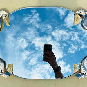 May include: Oval mirror with a reflective surface showing a blue sky with clouds. The mirror has a silver-colored frame with decorative, curved accents and gold-colored mounting hardware.