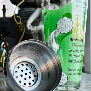 May include: A stainless steel cocktail shaker with a glass featuring a green label with a golf ball design and the words "Hole-in-One" and a cocktail recipe. The shaker is open, revealing its interior.