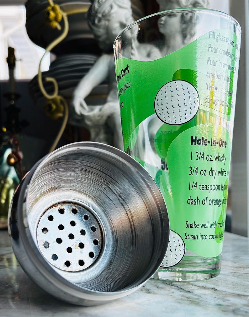 May include: A stainless steel cocktail shaker with a glass featuring a green label with a golf ball design and the words "Hole-in-One" and a cocktail recipe. The shaker is open, revealing its interior.
