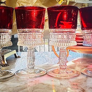 May include: Four antique wine glasses with ruby red bowls and clear, ornate stems. The glasses have a classic design with a wide base and a detailed, textured pattern on the stem and bowl base. The glasses are displayed on a marble surface.