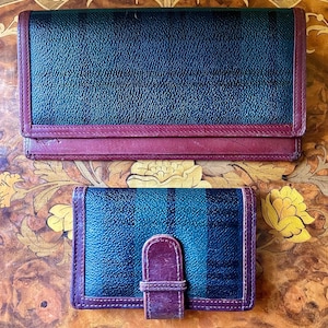 May include: Two leather wallets. The larger wallet is rectangular with a dark green and navy blue striped pattern and burgundy trim. The smaller wallet has a similar pattern and a burgundy strap closure.