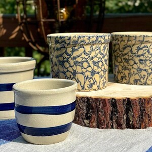 May include: Four ceramic crocks in a rustic setting. Two crocks have a blue and white pattern, while the other two have blue stripes. The crocks are displayed on a wooden slice and a blue and white tablecloth.