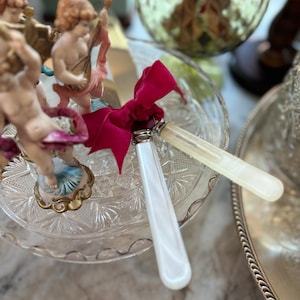 May include: Two porcelain cherub figurines with wings, holding musical instruments, are displayed on a clear glass cake stand with a red ribbon and two white handled cake servers.