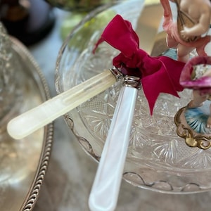 May include: Two decorative butter knives with white handles and silver accents are tied together with a deep red velvet ribbon. The knives rest on a clear, patterned glass plate, suggesting a vintage or antique aesthetic.