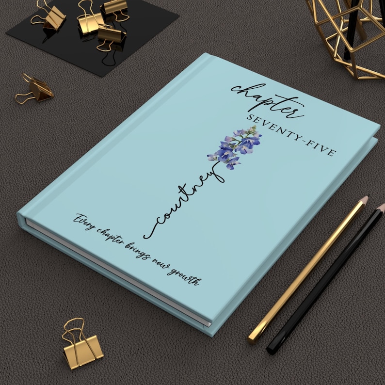 May include: A light blue hardcover journal featuring the text "chapter SEVENTY-FIVE", the name "courtney" written in script, a blue floral illustration, and the phrase "Every chapter brings new growth" printed on the front cover.