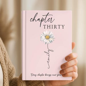 May include: A light pink hardcover journal featuring the text "chapter THIRTY" at the top, a central illustration of a white daisy with a yellow center, the name "emily" written in cursive script, and the text "Every chapter brings new growth" below.