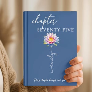 May include: A blue hardcover journal featuring the text chapter SEVENTY-FIVE at the top, a colorful lotus flower illustration in the center, the name Emily written in white script below the flower, and the text Every chapter brings new gro at the bottom.