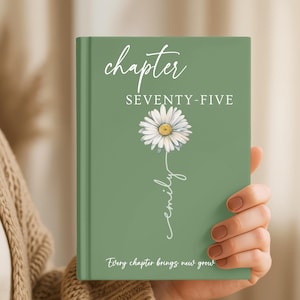 May include: A sage green hardcover book featuring the white text chapter SEVENTY-FIVE, a white daisy illustration with a yellow center, the name emily written in a cursive script, and the text Every chapter brings new growth at the bottom.