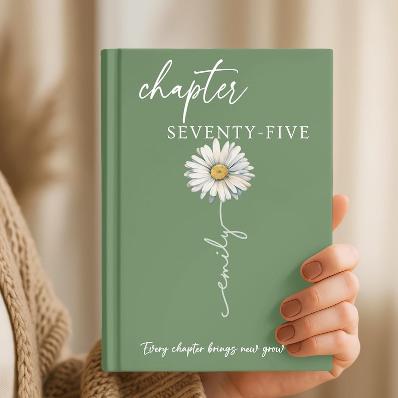 May include: A sage green hardcover book featuring the white text chapter SEVENTY-FIVE, a white daisy illustration with a yellow center, the name emily written in a cursive script, and the text Every chapter brings new growth at the bottom.