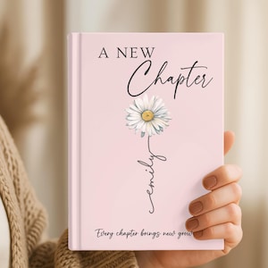 May include: A light pink hardcover journal featuring the text A NEW Chapter in black, a white daisy illustration with a yellow center, the name emily written in cursive script, and the text Every chapter brings new growth at the bottom.