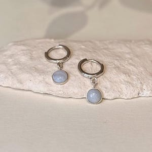 Op de afbeelding: Een paar zilveren oorringen met een kleine, ronde, lichtblauwe edelsteen hanger. De oorbellen worden gepresenteerd op een getextureerd, gebroken witte ondergrond. De edelstenen hebben een subtiele, doorschijnende kwaliteit.