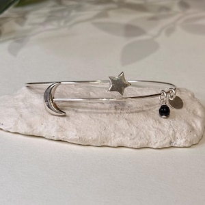 Op de afbeelding: Een zilveren armband met hemelse bedels. De armband heeft een halve maan en een ster, met een kleine zwarte kraal en een ronde bedel die aan de onderkant bungelt. De armband wordt gepresenteerd op een wit, getextureerd oppervlak.
