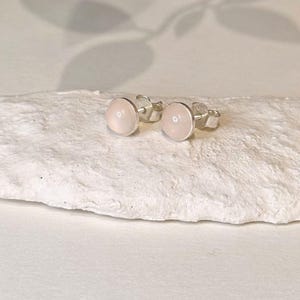 Può includere: Un paio di orecchini a bottone con una pietra rosa chiaro, circolare, incastonata in metallo argentato. Gli orecchini sono esposti su una superficie bianca e strutturata. Lo sfondo è bianco tenue con ombre di foglie.