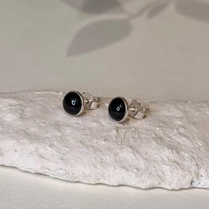 Puede incluir: Un par de pendientes de botón con una piedra negra redonda y brillante engastada en metal plateado. Los pendientes se muestran sobre una superficie texturizada de color blanco roto, con sombras suaves en el fondo. Son pequeños y minimalistas.