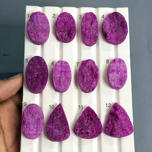 Puede incluir: Doce cabujones de piedra preciosa druzy de color magenta vibrante dispuestos en un tablero de exhibición blanco con ranuras. Las piedras están numeradas del uno al doce, con varias formas ovaladas y triangulares con superficies cristalinas.
