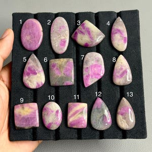 Pode incluir: Treze pedras preciosas polidas em cabochão dispostas numa bandeja de exibição preta com ranhuras. Cada pedra apresenta um padrão único de magenta vibrante e cinzento ou branco suave, numeradas de um a treze em texto branco.