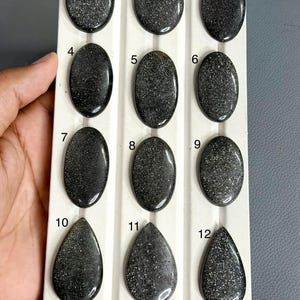 Può includere: Dodici cabochon in pietra del sole nera lucida disposti in una griglia su un vassoio espositore bianco. Le pietre sono numerate da uno a dodici, con nove forme ovali e tre a goccia, tutte caratterizzate da una superficie scintillante.