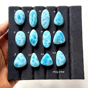 Puede incluir: Doce cabujones de larimar pulidos de diversas formas, incluyendo ovaladas, de pera y triangulares, están dispuestos en una bandeja de exhibición de espuma negra. Cada piedra presenta un patrón jaspeado azul cielo y blanco, numerado del 1 al 12.
