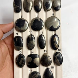May include: A collection of eighteen polished cabochon stones arranged on a white grooved display board. Each stone features a unique combination of metallic gold pyrite inclusions set against a dark black matrix, numbered from one to eighteen.