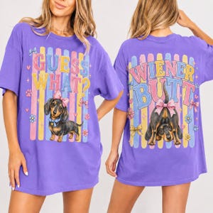 May include: A purple t-shirt with a dachshund graphic and the text "GUESS WHAT?" on the front. The back of the shirt features a dachshund graphic and the text "WIENER BUTT". The shirt has a relaxed fit.