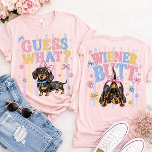 May include: Two pink t-shirts with dachshund graphics and text. One shirt says "GUESS WHAT?" with a dog wearing a bow. The other says "WIENER BUTT" with a dog's rear end. The shirts are next to jeans, sunglasses, and white sneakers.