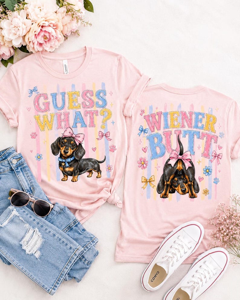 May include: Two pink t-shirts with dachshund graphics and text. One shirt says "GUESS WHAT?" with a dog wearing a bow. The other says "WIENER BUTT" with a dog's rear end. The shirts are next to jeans, sunglasses, and white sneakers.