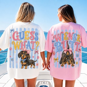 May include: Two oversized t-shirts, one white and one pink, with dog-themed graphics. The white shirt says "GUESS WHAT?" with a dachshund illustration. The pink shirt says "WIENER BUTT" with a dog's rear end.