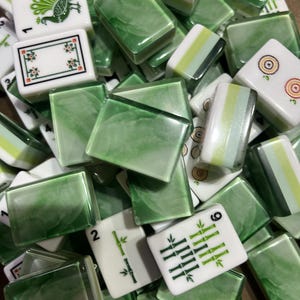 May include: A collection of rectangular mahjong tiles featuring a mix of solid white faces with green ink illustrations of bamboo, flowers, and birds, alongside translucent green marbled tiles with a smooth, glossy finish and rounded edges.