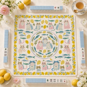 May include: A square mahjong mat featuring a repeating pattern of cats, lemons, and fish. The center displays a circular arrangement of three cats surrounded by fish and lemon slices. Four clear racks hold light blue and white mahjong tiles.