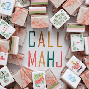 May include: A collection of Mahjong tiles with pink marble-patterned backs and white fronts featuring green, red, and blue floral and character designs, surrounding embroidered text that reads CALL MAHJ in green, gold, and red thread.