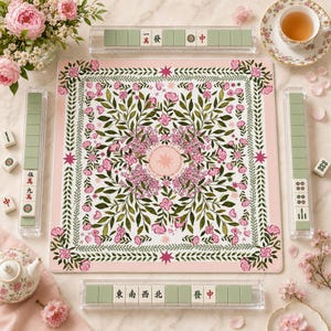 May include: A square mahjong mat featuring a white center with a symmetrical pattern of green leaves and pink flowers, bordered by a pink frame with floral motifs. Four clear acrylic racks hold mahjong tiles with Chinese characters and symbols.