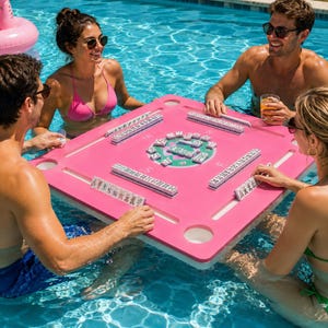 May include: A square, bright pink floating mahjong table designed for use in a swimming pool. The table features four integrated cup holders, recessed channels for tile racks, and a central circular area displaying mahjong tiles in play.