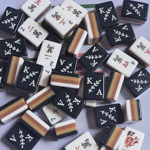 Könnte beinhalten: Eine Sammlung rechteckiger Mahjong-Spielsteine, bestehend aus schwarzen Steinen mit weißem Buchstaben K und Blattmotiv, weißen Steinen mit roten chinesischen Schriftzeichen sowie Steinen mit horizontalen Streifen in Schwarz, Braun und Weiß.