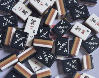 May include: A collection of rectangular Mahjong tiles featuring black tiles with white letter K and a leaf design, white tiles with red Chinese characters, and tiles with horizontal stripes in black, brown, and white colors.