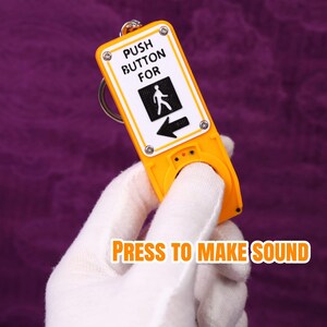 May include: A yellow keychain shaped like a pedestrian crossing button. The white sign reads "PUSH BUTTON FOR" with a pedestrian symbol and an arrow. The text "PRESS TO MAKE SOUND" is also visible.