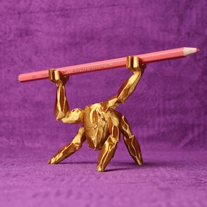 Peut inclure: Une sculpture dorée de style origami représentant une personne tenant un crayon rose. Le crayon porte l'inscription "SW CLASSIC COLOUR" et "STEEL 131". La sculpture est sur un fond violet.