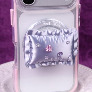May include: A light purple phone grip in the shape of a pillow, adorned with multiple rhinestones. The pillow grip is attached to a clear phone case with pink accents. The rhinestones are clear, pink, and purple.