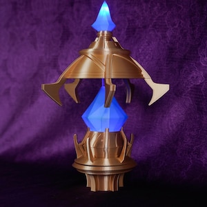 May include: A gold-coloured, ornate decorative object with a blue, glowing crystal at its centre. The object has a multi-tiered design with pointed and curved elements. The top and bottom sections are circular, and the crystal is diamond-shaped.