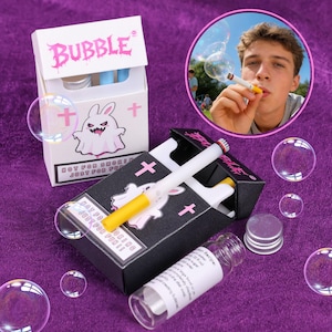 Puede incluir: Un juego de artículos novedosos que presenta una caja blanca y una caja negra, ambas diseñadas para parecerse a paquetes de cigarrillos. La caja blanca está etiquetada "BUBBLE" en rosa, y la caja negra tiene el texto "NOT FOR SMOKING JUST FOR FUN!!" El juego incluye cigarrillos para hacer burbujas.