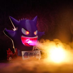 Puede incluir: Una figura de Gengar morado con ojos y boca rojos brillantes, que emite una luz rosa-púrpura. La figura tiene una sonrisa amenazante y está rodeada de humo. Un pequeño letrero con texto japonés e ilustraciones de personajes está al frente.