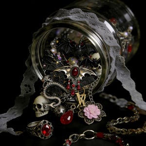 Puede incluir: Un tarro de cristal rebosante de joyas y accesorios góticos. La colección incluye un colgante de murciélago con una gema roja, abalorios de calaveras y un anillo con una gran piedra roja. Una cinta de encaje gris decora el tarro.