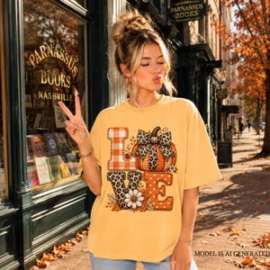 May include: A mustard yellow t-shirt featuring a graphic print of the word LOVE. The letter L has an orange plaid pattern, the O is a pumpkin with a leopard print bow, the V has a leopard print, and the E is orange with white polka dots.