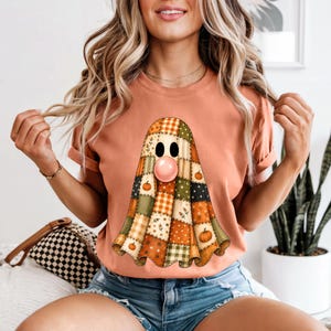 Może przedstawiać: Brzoskwiniowy t-shirt z krótkim rękawem z centralną grafiką ducha wykonanego z patchworkowego wzoru w odcieniach pomarańczu, zieleni i kremu. Duch ma dwoje czarnych owalnych oczu i różową bańkę z gumy do żucia pośrodku.