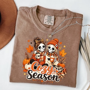 Puede incluir: Una camiseta marrón de manga corta con un gráfico de dos esqueletos con bufandas y gorros que sostienen tazas. El diseño incluye calabazas, hojas de otoño y el texto "Cozy Season" en letras de color naranja y blanco.