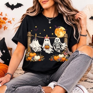 May include: A black short-sleeve t-shirt featuring a graphic of three ghosts hanging from a clothesline between two wooden posts. The ghosts are wearing a witch hat, a pink bow, and a skeleton costume, surrounded by pumpkins, flowers, and a moon.