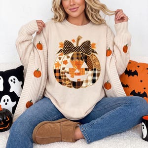 Könnte beinhalten: Ein beiges Langarm-Sweatshirt mit einer großen zentralen Kürbisgrafik, die aus verschiedenen karierten und blattgemusterten Stoffstücken besteht und von einer schwarz-weißen gepunkteten Schleife gekrönt wird. Die Ärmel sind mit kleinen orangefarbenen Kürbissen bestickt.