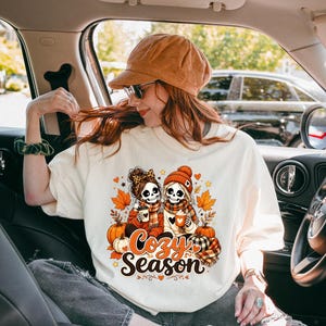 May include: An off-white t-shirt featuring a graphic of two skeletons wearing hats and scarves, holding mugs. The design includes orange pumpkins, autumn leaves, and the text "Cozy Season" in a brown script font, surrounded by small heart accents.