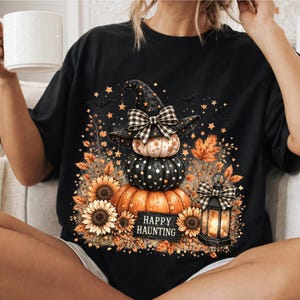 May include: A black t-shirt featuring a graphic print of three stacked pumpkins, with the top pumpkin wearing a black witch hat adorned with a plaid bow. The design includes sunflowers, autumn leaves, a lit lantern, and the text HAPPY HAUNTING.